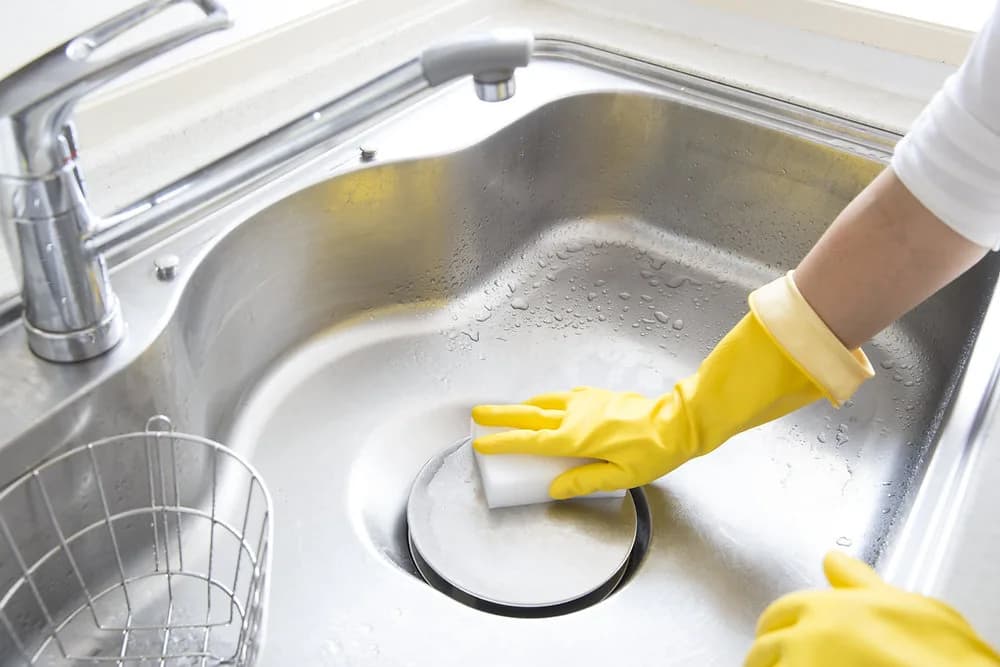 Hand in gelbem Haushaltshandschuh reinigt mit einem Schwamm den Ablauf einer Edelstahl-Küchenspüle