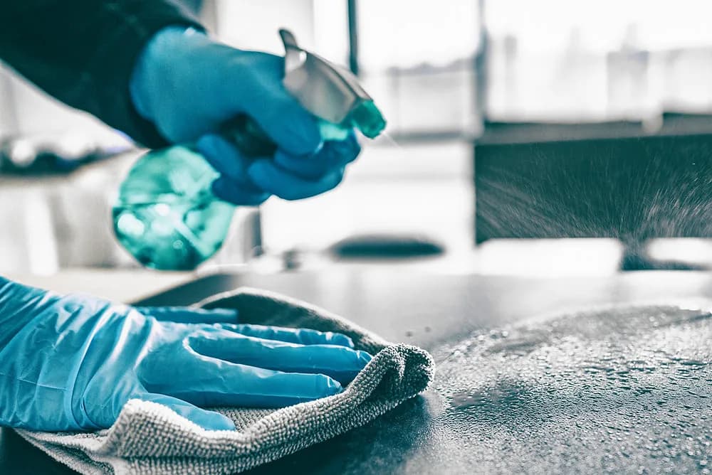 Hand in blauem Schutzhandschuh sprüht Reiniger auf eine dunkle Arbeitsplatte, die zweite Hand wischt mit einem Mikrofasertuch nach