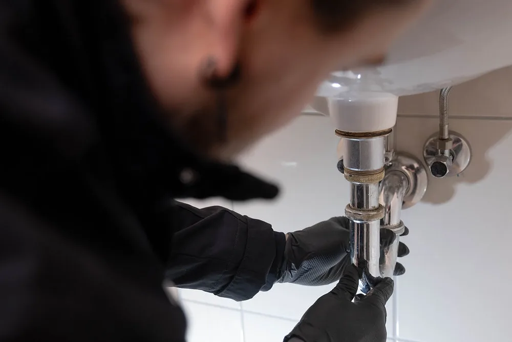 Handwerker in schwarzen Arbeitshandschuhen löst den verchromten Siphon unter einem Waschbecken im gefliesten Bad