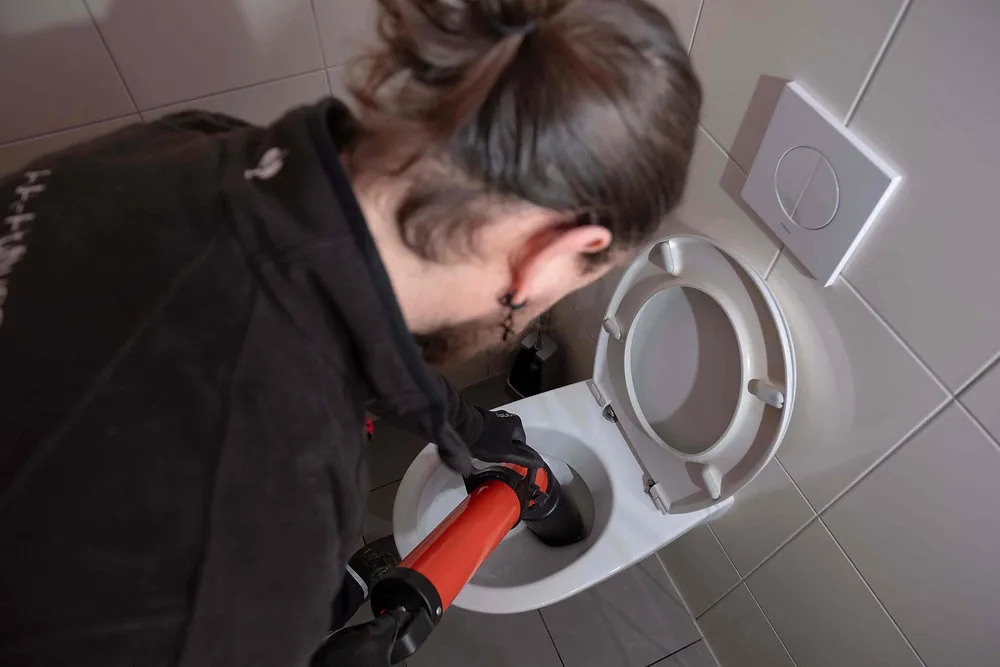 Monteur setzt eine rote Rohrreinigungspumpe am Ablauf einer Toilette an
