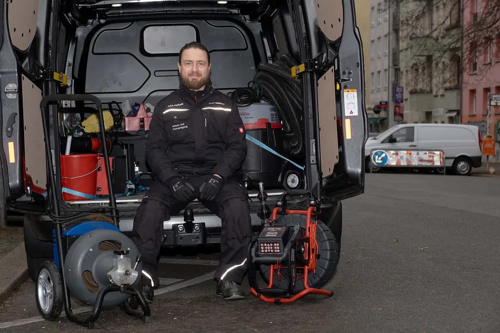 Inhaber von AAA-Abfluss sitzt in der geöffneten Heckklappe seines Einsatzfahrzeugs, davor Rohrreinigungsspirale und Kameratechnik