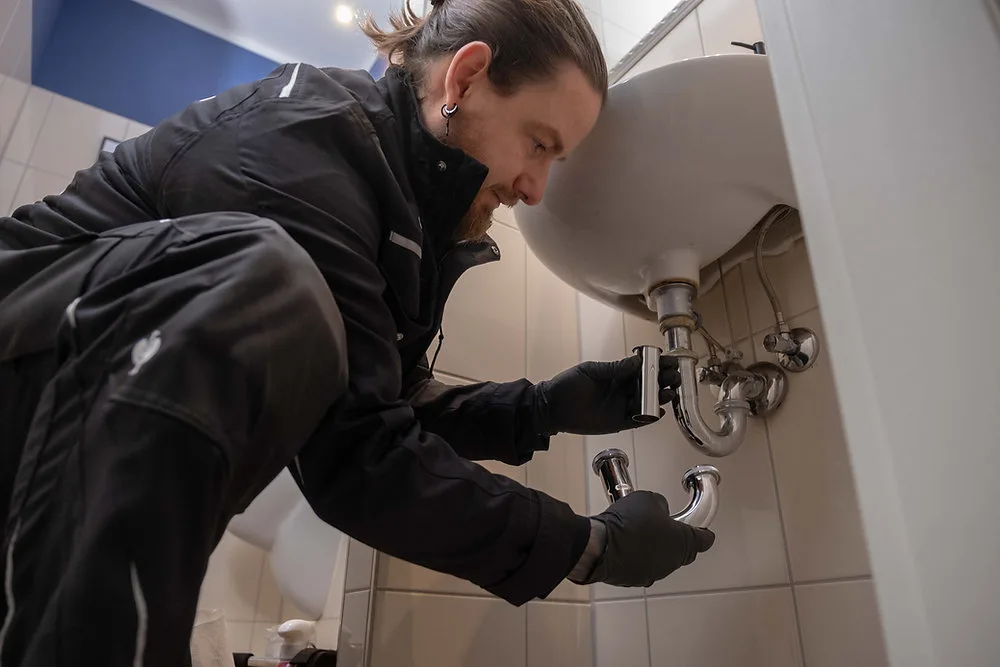 Monteur beugt sich unter ein Waschbecken und arbeitet mit beiden Händen am Siphon der Ablaufleitung